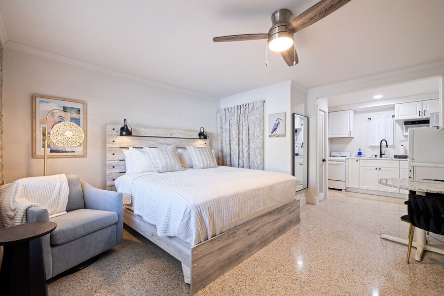 A spacious sleeping area features a king-size bed with a light-colored bedspread. A ceiling fan hangs above, providing airflow. A comfortable chair is placed in the corner, and a kitchenette is visible in the background, equipped with modern appliances.