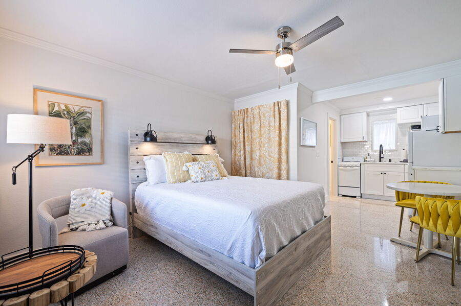 The living space features a light-colored queen size bed with decorative pillows, accompanied by a comfortable armchair and side table. A ceiling fan is present above, and a kitchenette is visible in the background. A dining table with two vibrant yellow chairs is situated nearby.