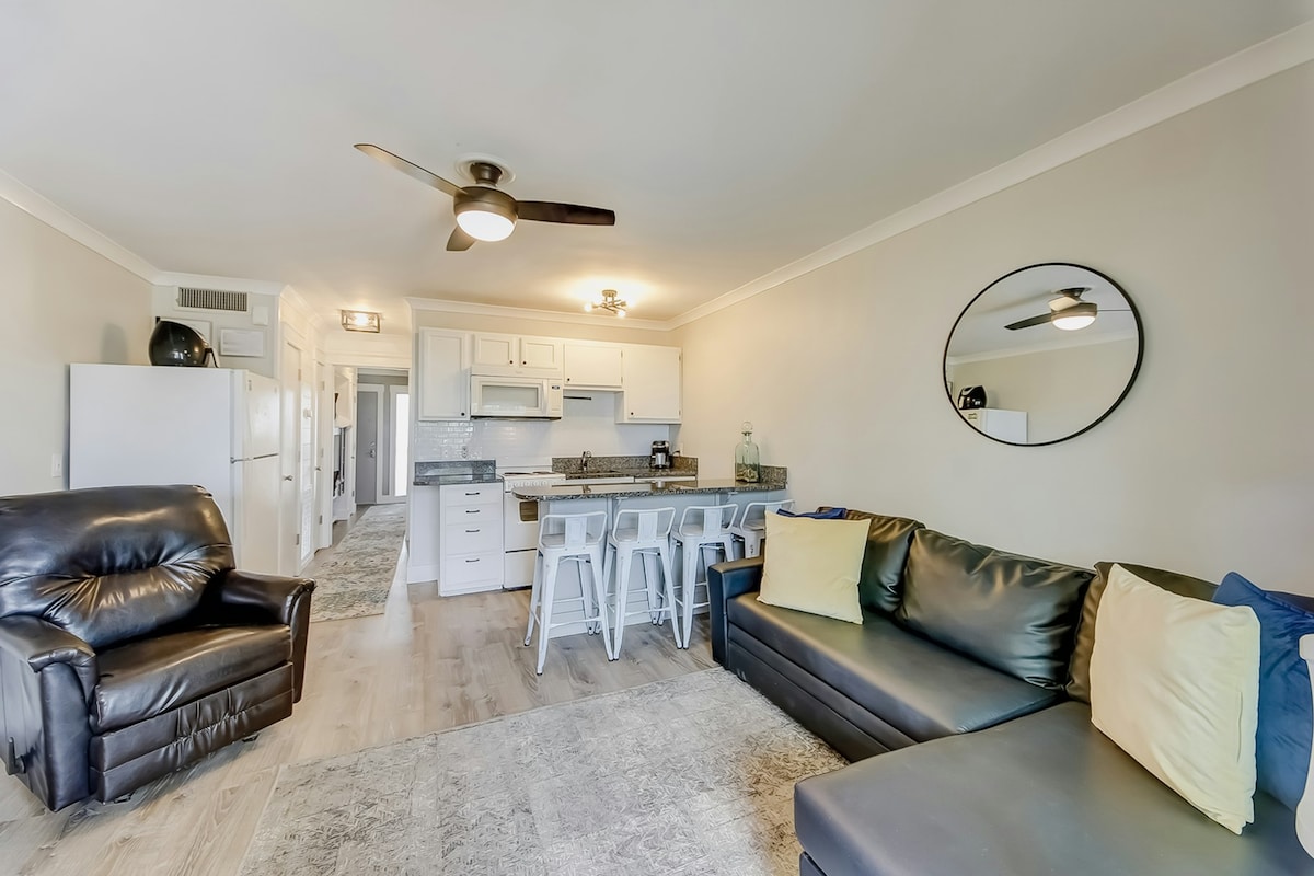 A spacious living area features a dark leather sectional sofa with colorful decorative pillows and a black armchair. A modern kitchen with white cabinetry and a breakfast bar is visible, complemented by a circular mirror reflecting the open space.