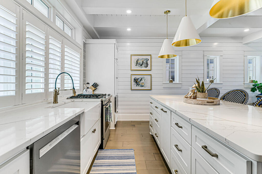 The kitchen features bright white cabinetry and a large island with a marble countertop. Natural light filters through plantation shutters on the windows, highlighting the stainless-steel appliances, including a gas range. Decorative wall art and a centerpiece plant add a touch of coastal style.