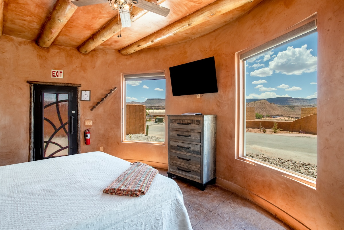 A spacious sleeping area features a king bed with a white quilt and a cozy accent throw. Large windows allow natural light to flood the room, offering scenic views of the surrounding landscape. A wall-mounted TV provides modern entertainment options.