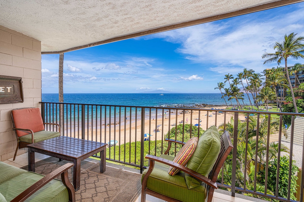 A beach-front lanai offers expansive views of the ocean, featuring gentle waves and sunlit sands below. Comfortable seating with colorful cushions is positioned around a wooden table. Lush greenery and swaying palm trees frame the scene, enhancing the coastal ambiance of this retreat.