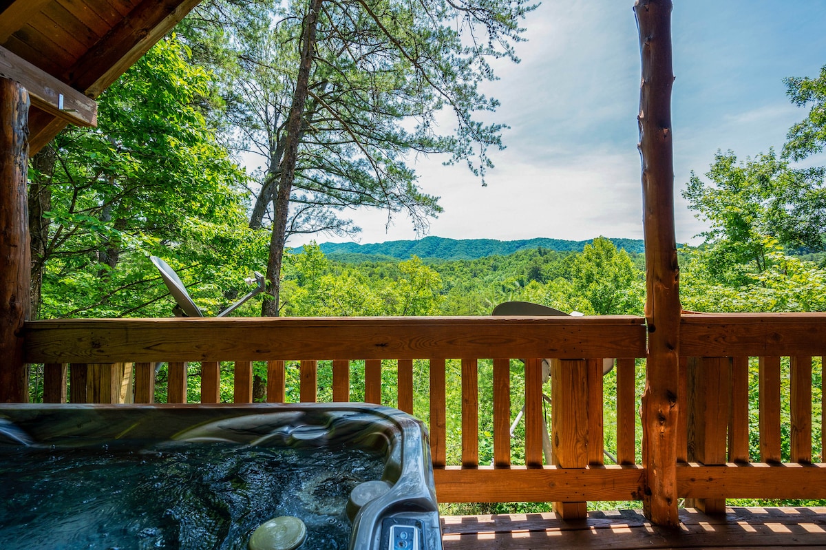 An outdoor deck features a large hot tub overlooking a lush green landscape, showcasing rolling hills and trees. The railing of the deck highlights the serene view of the Smoky Mountains, inviting moments of relaxation amid nature.