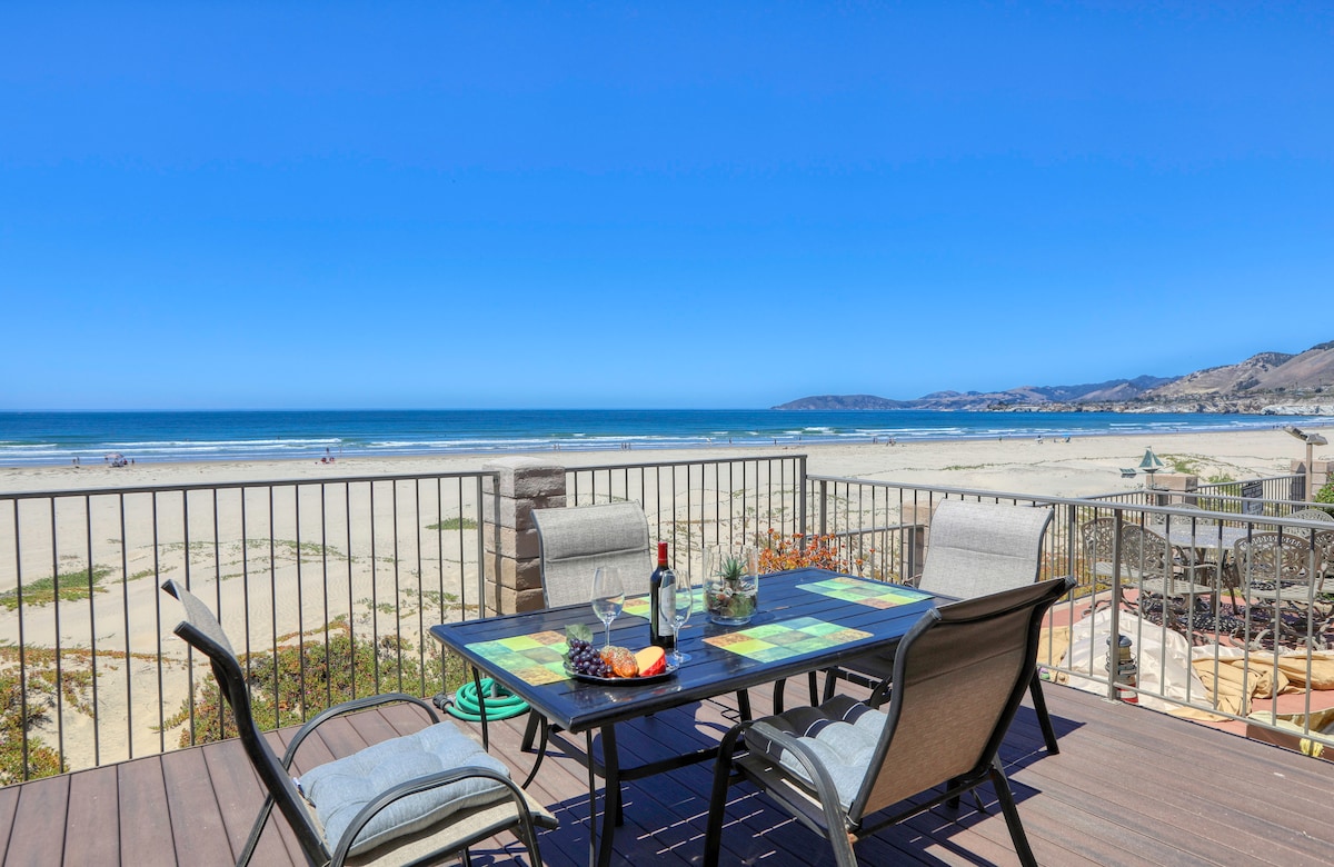 An oceanfront deck features a dining table with four chairs, accompanied by plates and glasses. The expansive view showcases the sandy beach and the Pacific Ocean under a clear blue sky, providing a serene backdrop for outdoor dining.