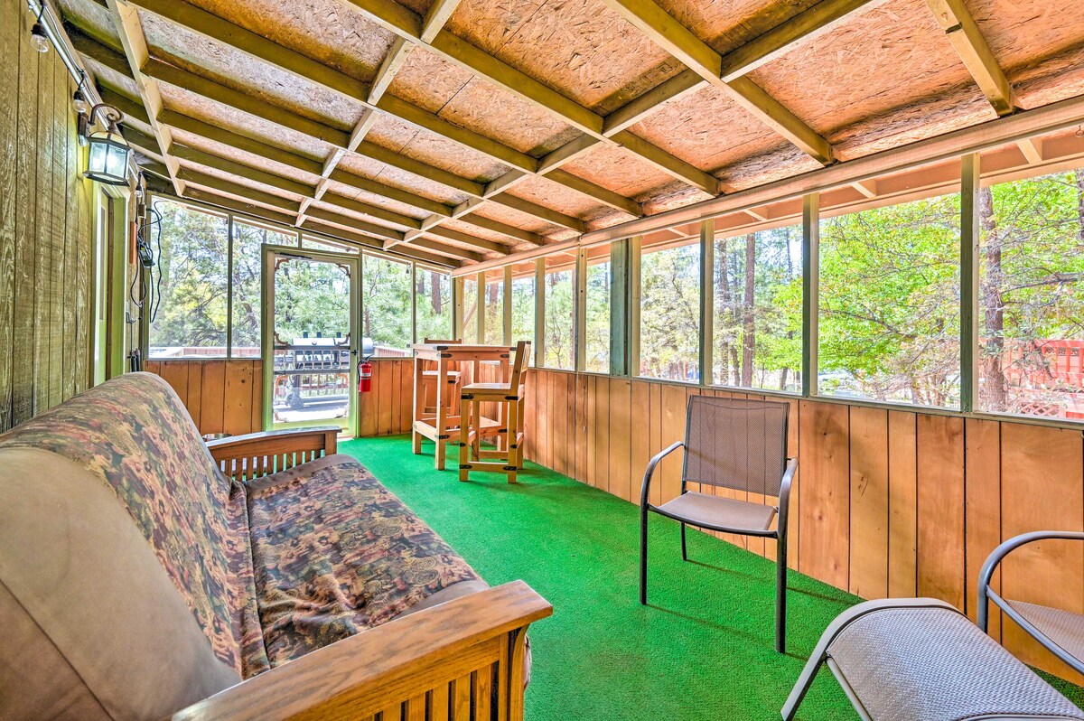 A screened patio is presented with a wooden couch adorned with patterned upholstery and a light wooden table. Chairs are arranged around the area, with natural light filtering through the windows, providing a view of the surrounding greenery.