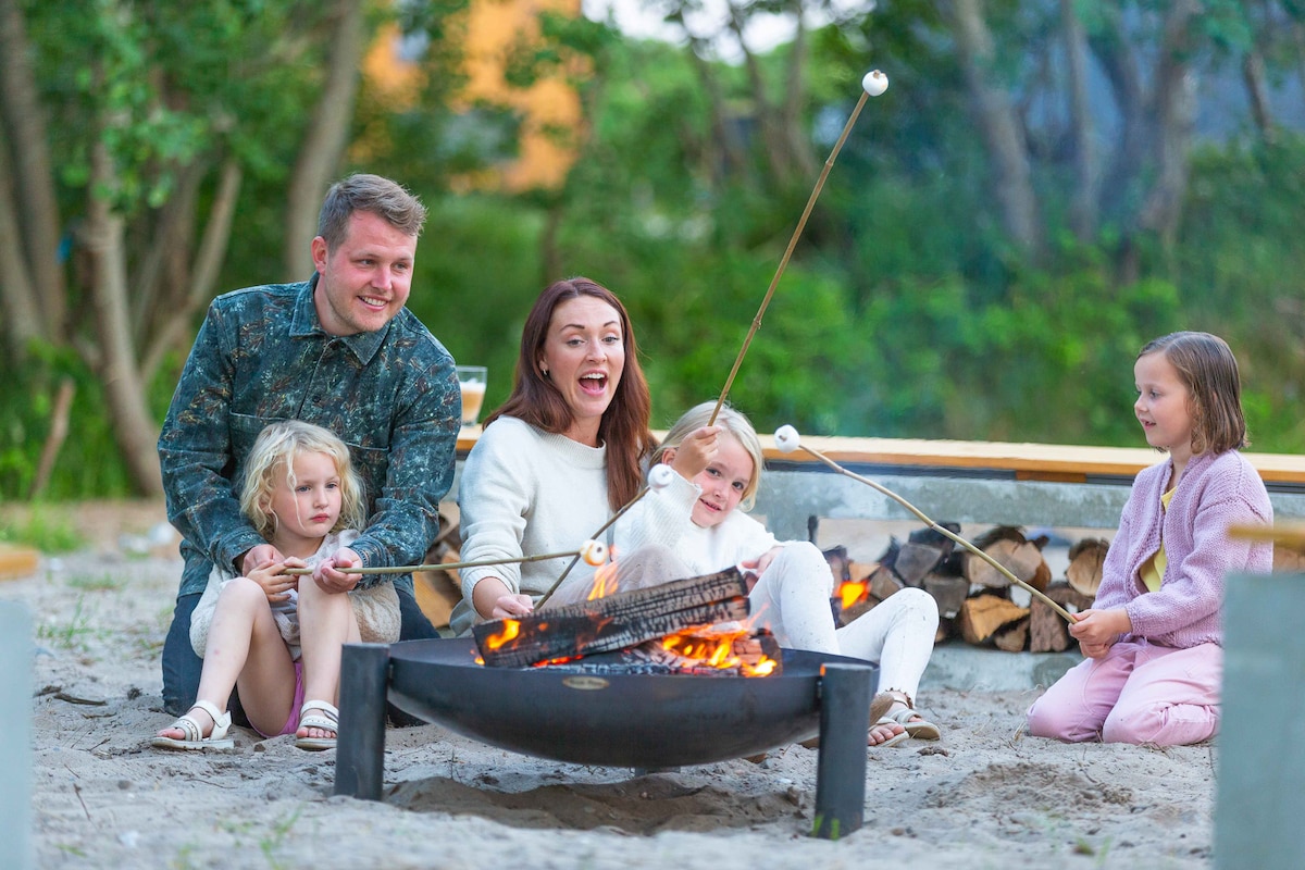 A family gathers around a fire pit in a sandy area, with logs stacked nearby. Each member holds a skewer for roasting marshmallows over the flames. Surrounding greenery enhances the natural setting, creating a relaxed atmosphere for evening gatherings.