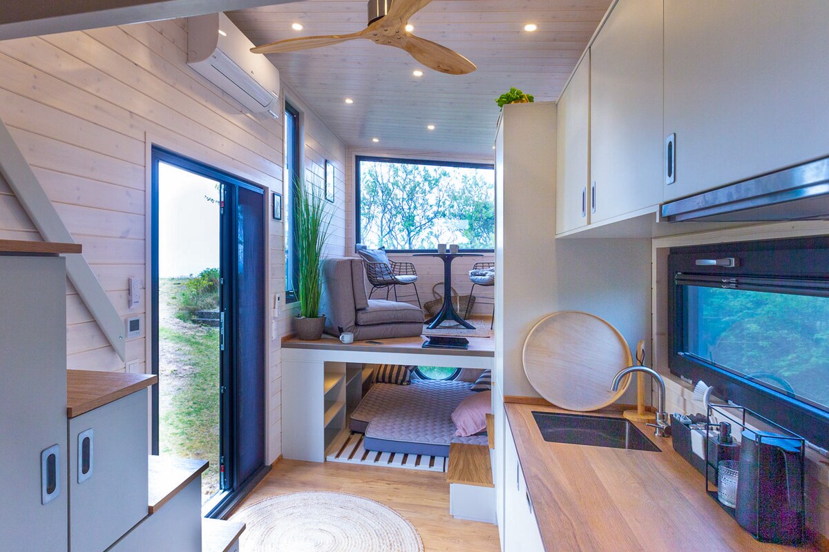 An open interior layout showcases a light-filled space with large windows offering views of the outdoors. Minimalist furnishings include a comfortable seating area and a well-equipped kitchen. Light wooden accents and a smooth floor create a warm ambiance, complemented by a ceiling fan for airflow.