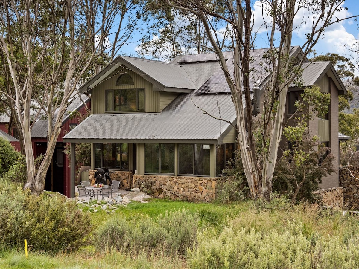 A spacious chalet is surrounded by native bushland, featuring large windows that invite natural light. A stone facade complements the modern design, and outdoor seating is visible on the patio, providing an inviting area for relaxation amidst the natural setting.