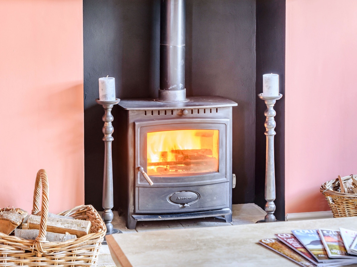 A woodburner is centrally placed within a black surround. It glows warmly with flickering flames, complemented by two decorative candlesticks on either side. A wicker basket filled with logs is positioned nearby, enhancing the cozy atmosphere of the living space.