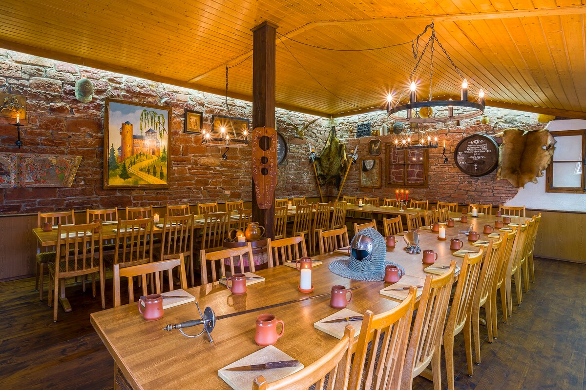 A spacious dining area is filled with wooden tables and chairs, arranged to accommodate large groups. Rustic stone walls and wooden beams enhance the warm ambiance. Lighting fixtures hang from the ceiling, casting a soft glow over decorative elements and place settings around the tables.