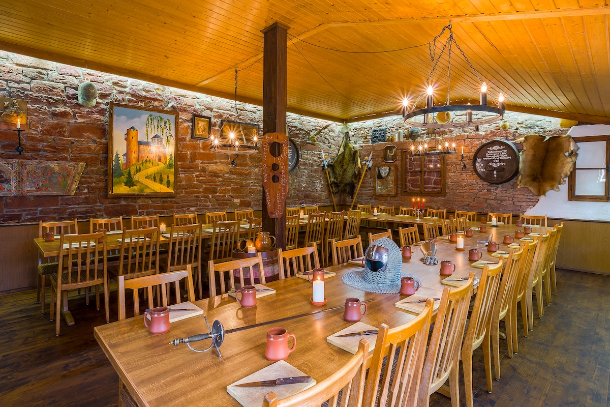A spacious dining area features multiple wooden tables arranged with chairs, ready for guests. The walls are adorned with rustic decor, including paintings and artifacts, while warm lighting fixtures provide an inviting glow across the space.