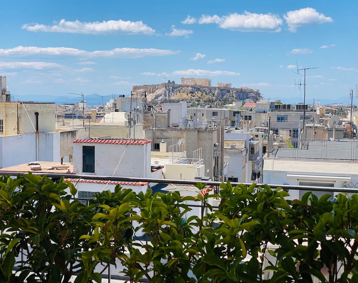 A panoramic view of the Acropolis is captured from a private terrace, framed by greenery. The vibrant blue sky and scattered clouds provide a serene backdrop to the historical site, while rooftops of neighboring buildings create a contrasting urban landscape.