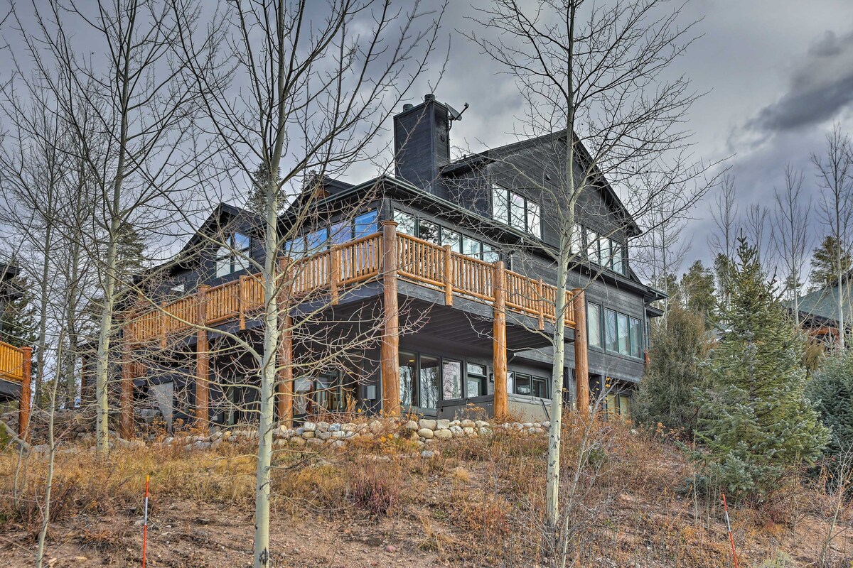 A modern mountain retreat is set against a cloudy sky, showcasing a spacious wrap-around deck with wooden railings. The exterior features dark wood siding and large glass windows, allowing views into the interior. Surrounding trees and natural landscaping enhance the home’s mountain setting.