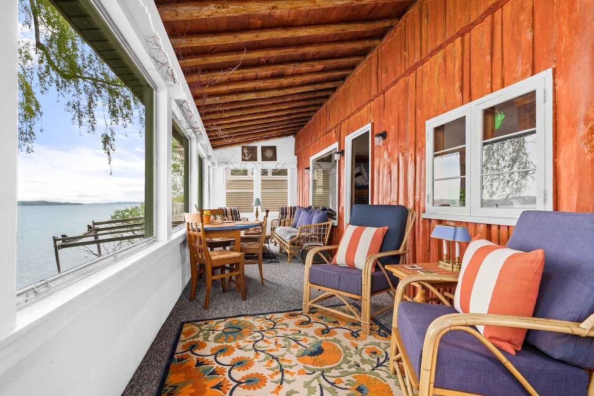 A welcoming 3-season room features comfortable seating, including two chairs adorned with bright pillows. A dining table is positioned nearby, with a patterned area rug underfoot. Large windows frame the serene views of Suttons Bay, while wooden beams add a touch of rustic charm.