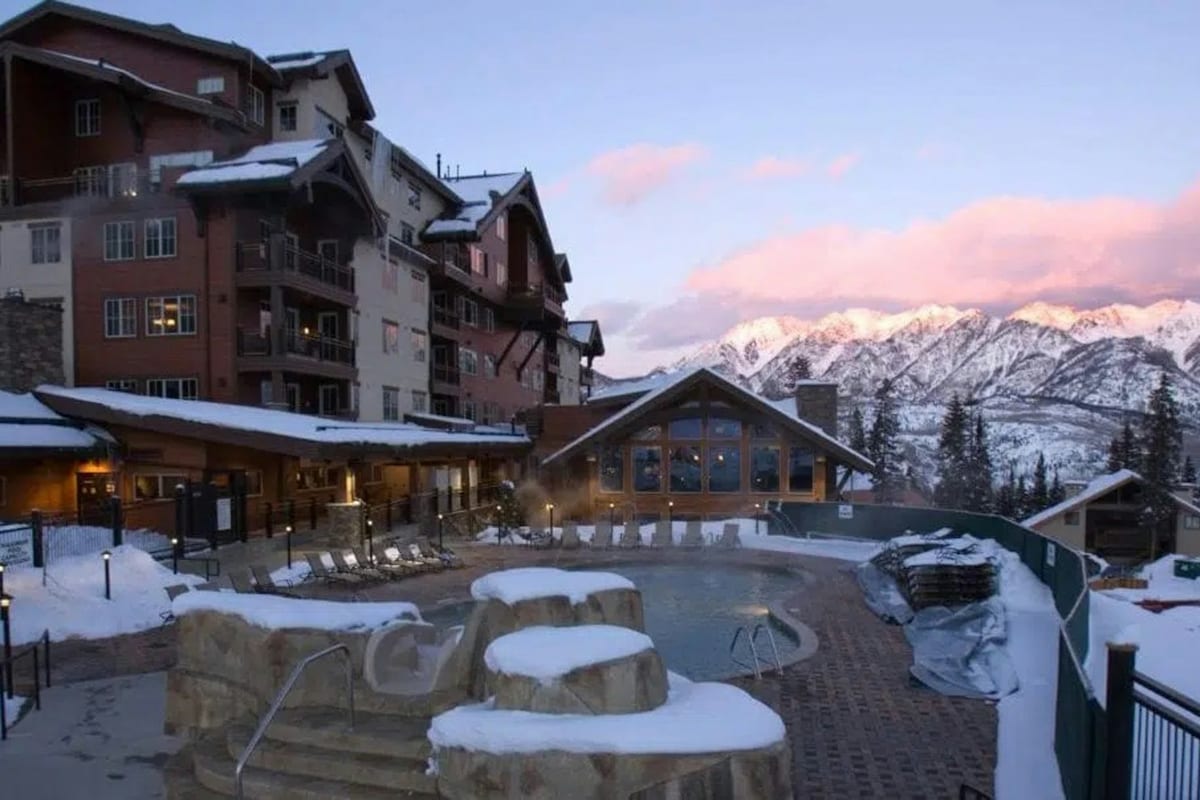 A spacious outdoor area is presented, featuring a heated pool surrounded by a stone edge. Lounging chairs are placed nearby, with majestic mountain views in the background. The sky displays soft pink hues as dusk approaches, creating a serene atmosphere.