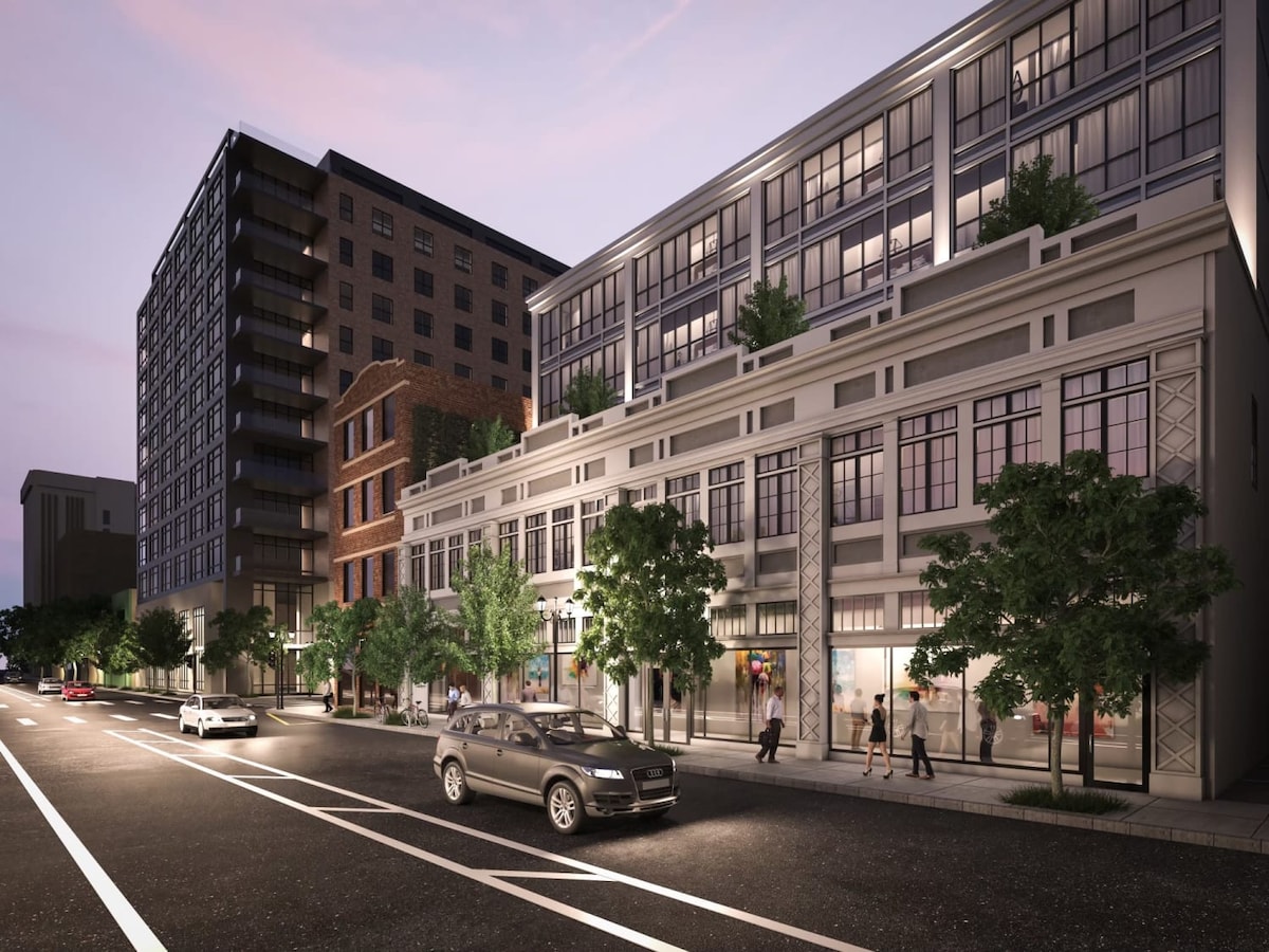 The exterior view showcases a modern multi-story building, blending contemporary and historic architectural elements. Lush greenery frames the entrance, and large windows provide an inviting appearance. Pedestrians stroll along the sidewalk, and vehicles are parked on the street, illustrating a vibrant urban setting.