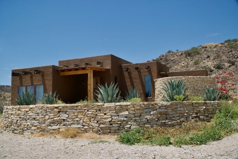 The exterior features a rustic adobe-style casita surrounded by natural landscaping. Prominent agave plants are visible in the front, and large windows reflect the desert landscape. The structure is adorned with wooden accents and sits against a backdrop of rugged hills under a clear blue sky.