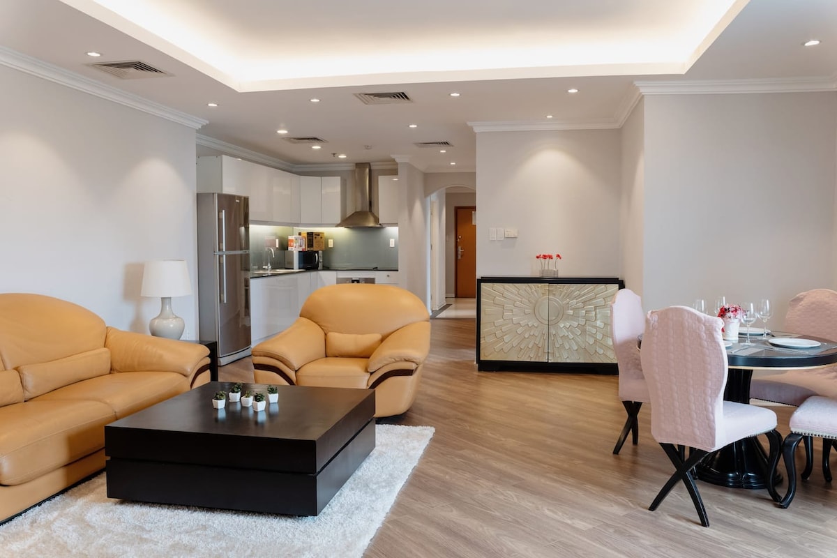 A spacious living area is highlighted by soft brown leather sofas and a dark coffee table. A bright dining set with pink chairs is positioned nearby, while a sleek, modern kitchen is visible in the background, featuring stainless steel appliances and soft lighting.