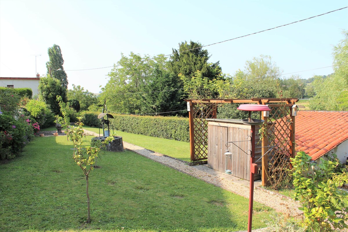 The garden area features well-maintained grass surrounded by vibrant greenery and hedges. A decorative wooden trellis stands near the garden with a small storage shed visible. Various trees and shrubs add to the serene outdoor setting, framed by gentle slopes of the countryside.