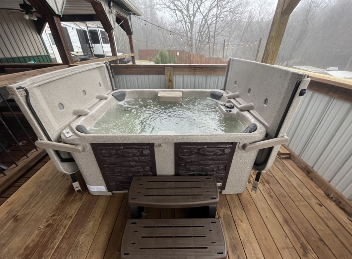 A spacious hot tub is situated on a wooden deck, featuring a clear water surface. Access is provided via two sturdy steps. Surrounding the tub, natural scenery creates a serene atmosphere, while the misty backdrop adds to the peaceful setting.