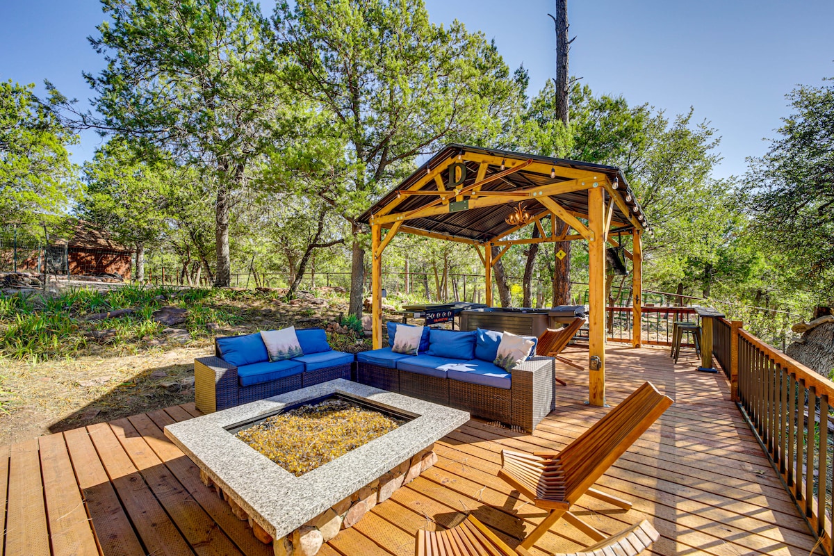 An outdoor seating area features a comfortable sectional sofa with blue cushions, positioned around a stone fire pit. A gazebo provides shade, and a wooden deck extends into a forested landscape, creating a serene space for relaxation.