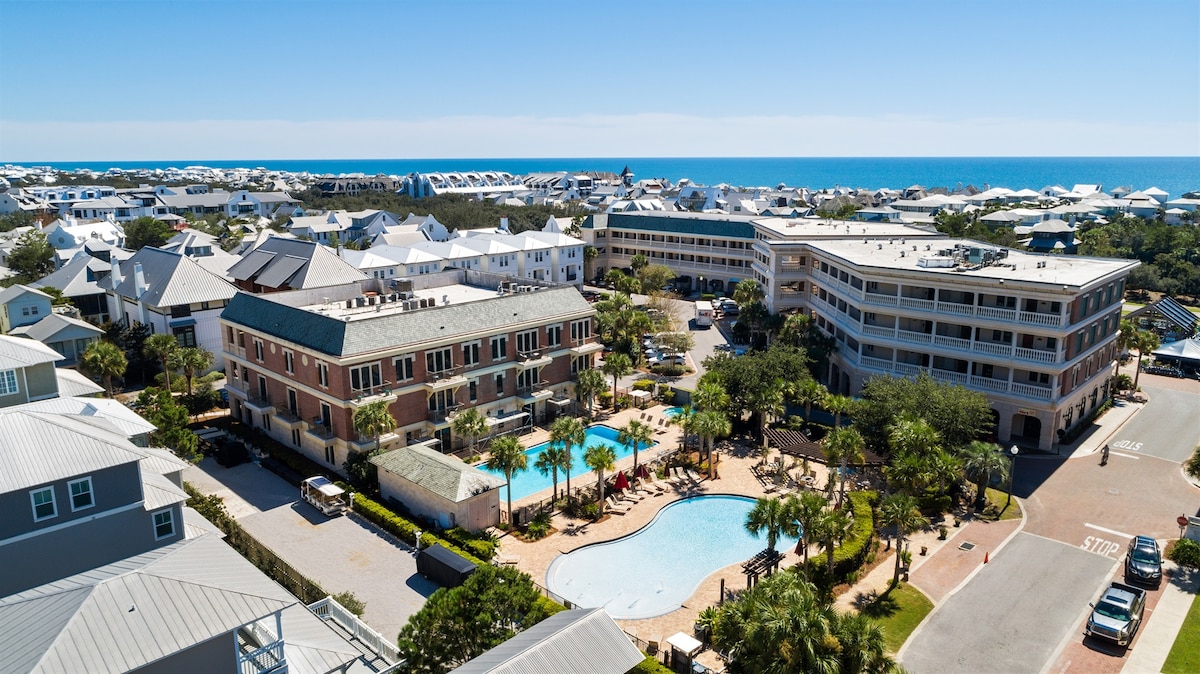 An aerial view captures the resort complex with its inviting pool area surrounded by palm trees. The ocean is visible in the distance, with rows of beach houses lining the horizon. Bright blue skies add to the serene coastal atmosphere.