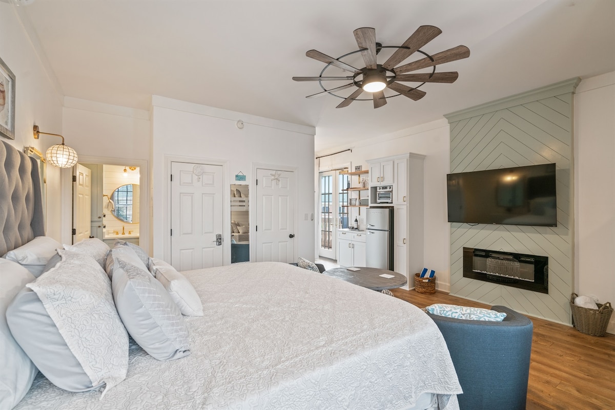 A spacious studio bedroom features a large bed with a textured white comforter and decorative pillows. A modern ceiling fan hangs above, and an electric fireplace is installed beneath the mounted television. A kitchenette and adjoining bathroom are visible in the background.
