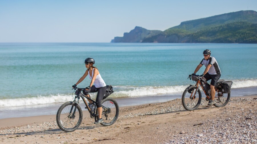 Two individuals are seen riding bicycles along a sandy beach. The ocean stretches out in the background, meeting a clear blue sky. The coastline features rolling hills, creating a serene backdrop for their ride.