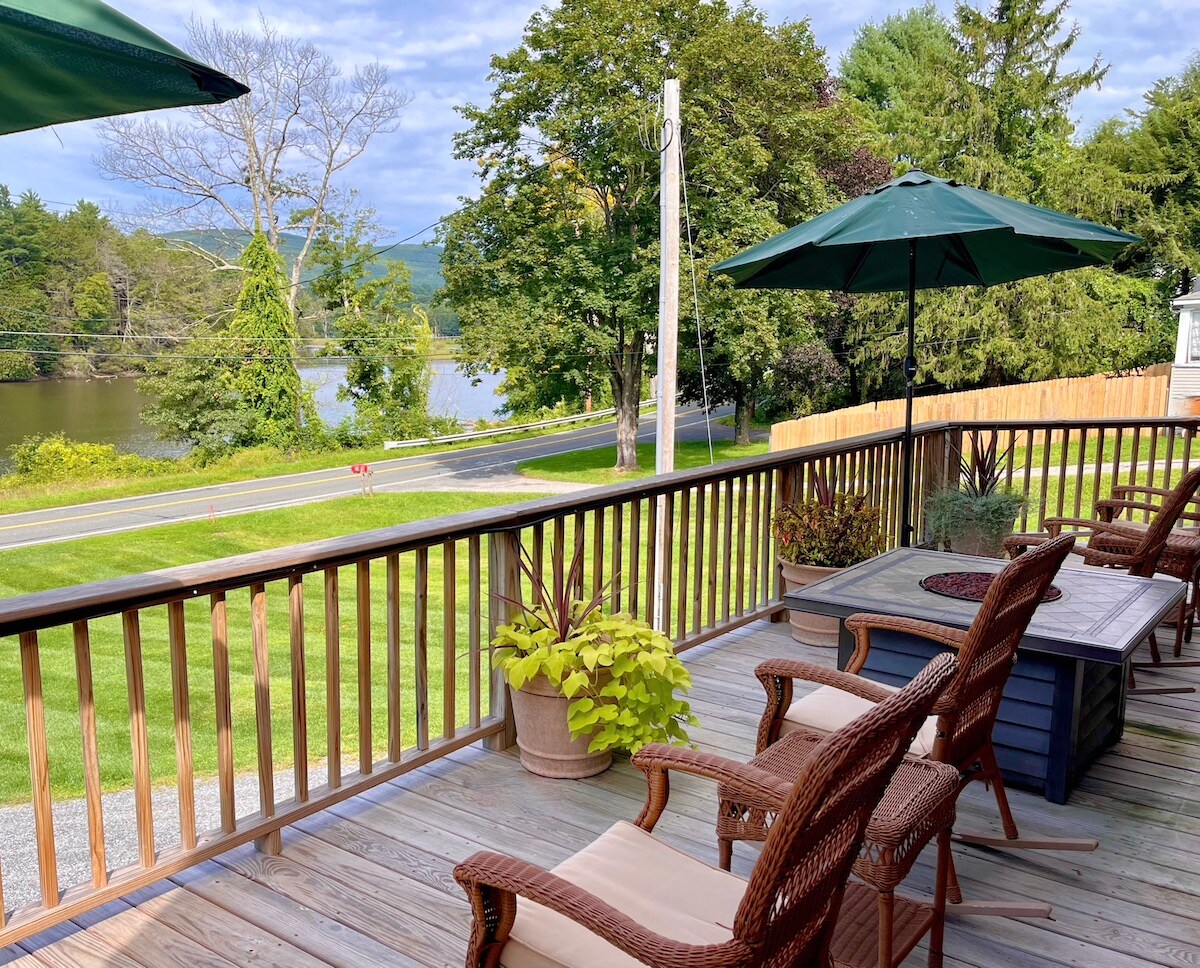 A wooden deck is visible, featuring several comfortable chairs and two green umbrellas providing shade. Potted plants add a touch of greenery. The Housatonic River is seen in the background, with trees lining the opposite shore and a scenic view of the surrounding landscape.