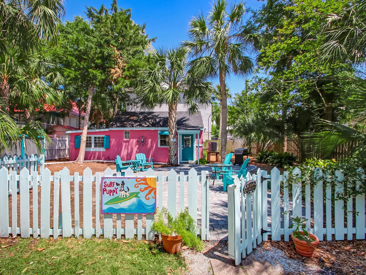 The exterior of the beach cottage is showcased, featuring a cheerful pink facade with aqua accents. A white picket fence surrounds the property, while palm trees and greenery add a tropical feel. Outdoor seating areas are visible on the paved patio.