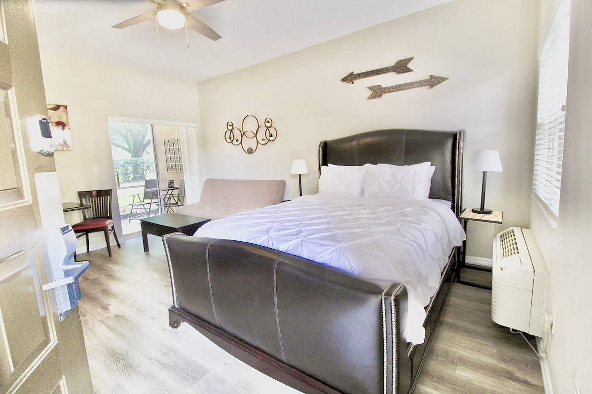 A spacious sleeping area features a large, dark leather bed with a plush white comforter. A futon couch is positioned nearby. Natural light enters through a window, framed by light curtains, with additional seating visible in the open layout leading to the patio.