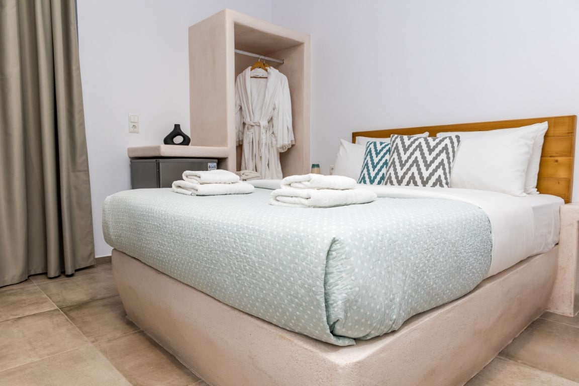 A well-appointed sleeping area features a comfortable double bed adorned with a light blue quilt. Neatly arranged towels are placed at the foot of the bed. A storage unit with a hanging robe and shelves is positioned nearby, providing practical amenities for guests.