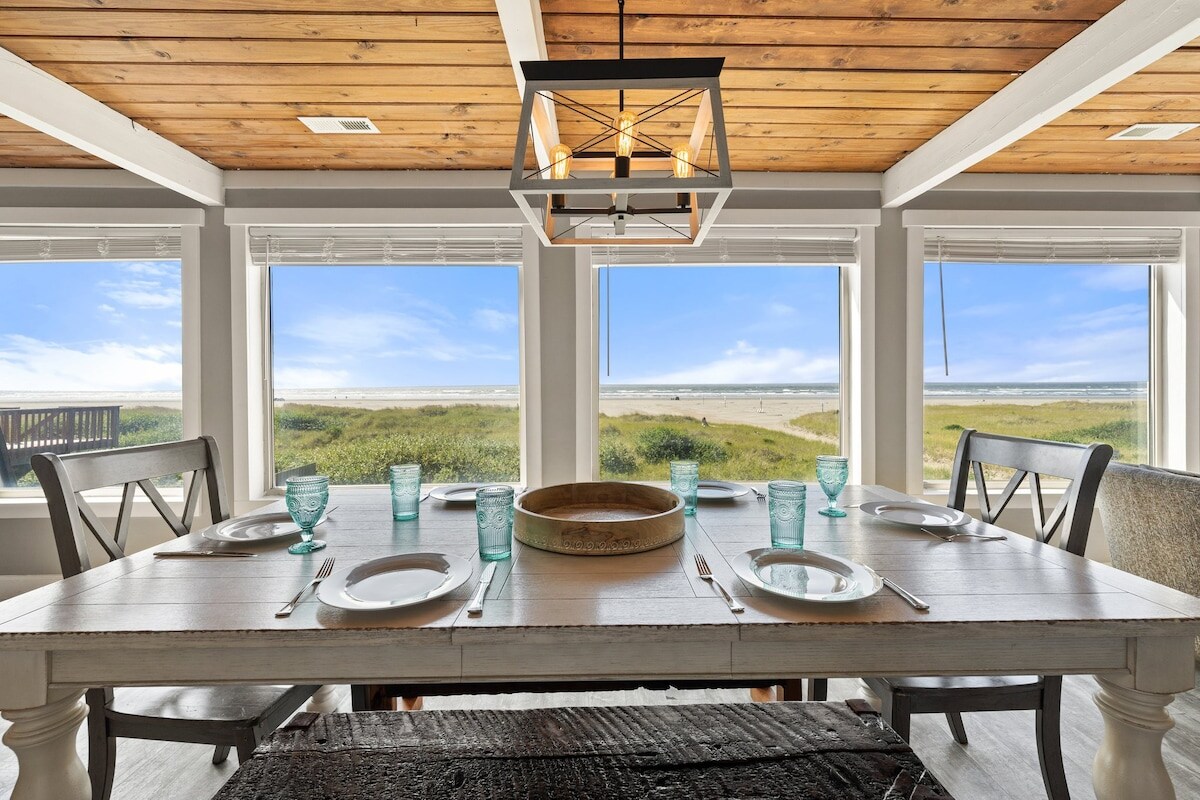A spacious dining area features a large table set for meals. Sunlight streams through multiple windows, showcasing views of the sandy beach and grassy dunes. Light wooden beams accent the ceiling, while the table is adorned with turquoise glasses and simple plates.