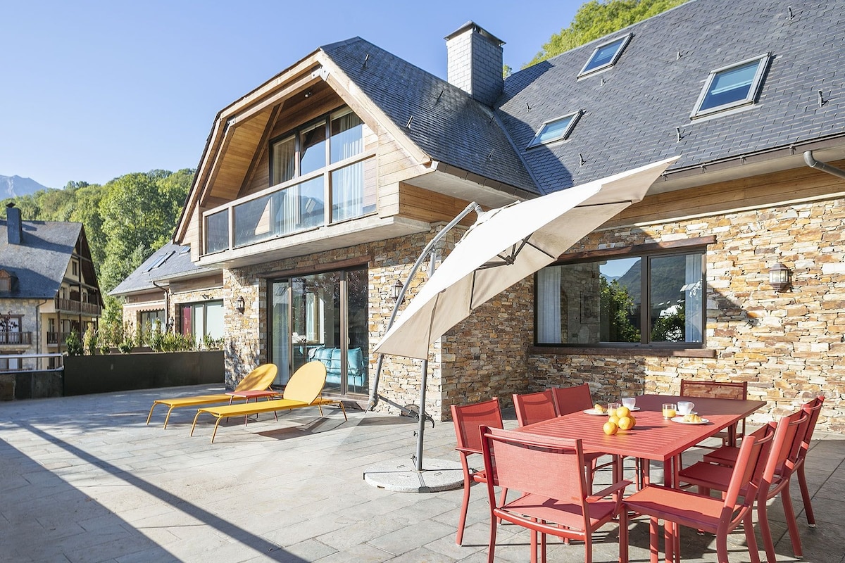 An outdoor terrace features a dining table surrounded by red chairs, with an umbrella providing shade. Two lounge chairs are positioned nearby on the stone flooring. The house, with wooden and stone elements, showcases large windows reflecting natural light against a backdrop of greenery.