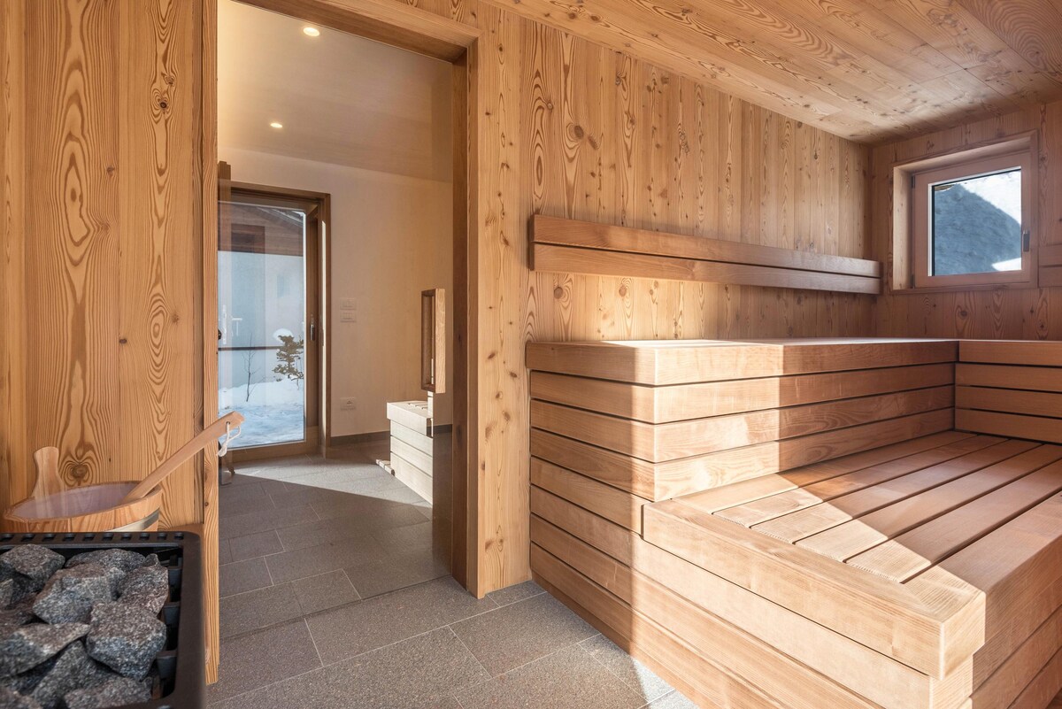 A wooden sauna is presented with smooth, warm-toned benches and stone features. Natural light filters through a small window, illuminating the space. A cozy chair is positioned nearby, creating a relaxing environment for guests.