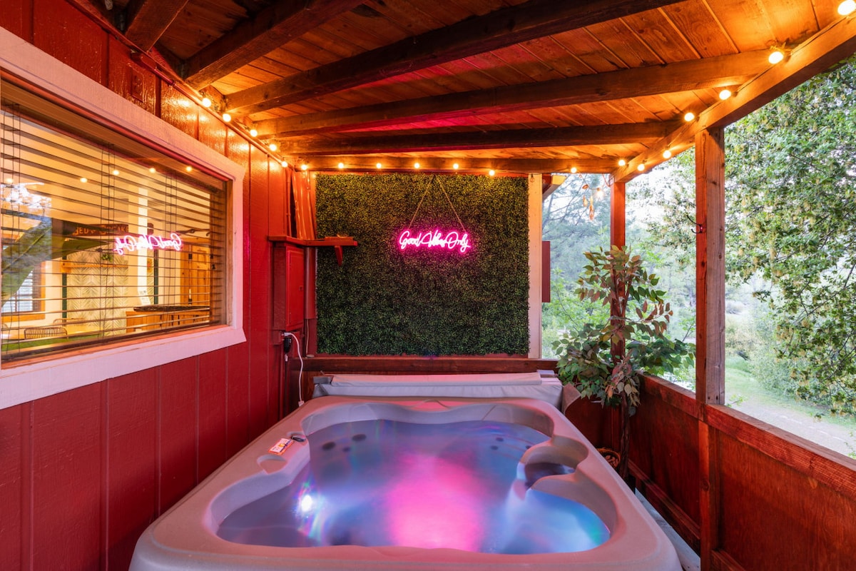 A private hot tub is nestled on a covered deck, surrounded by wooden beams and ambient lighting. A neon sign adds a pop of color, while greenery provides a natural backdrop. Large windows allow for views of the outdoors, enhancing the relaxing atmosphere.