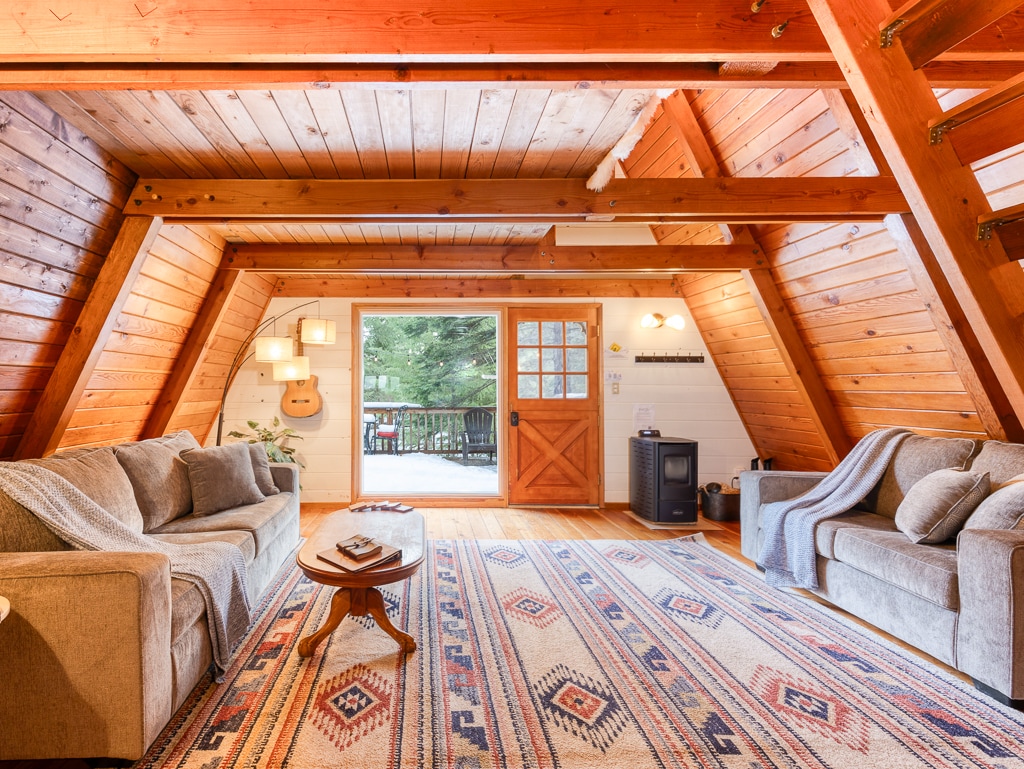 The inviting living area is adorned with wooden beams and features two comfortable sofas draped with light throws. A patterned area rug lies on a wooden floor, complementing a small round table. A door leads to the outside, framed by lush trees, and a pellet stove is positioned warmly in the corner.