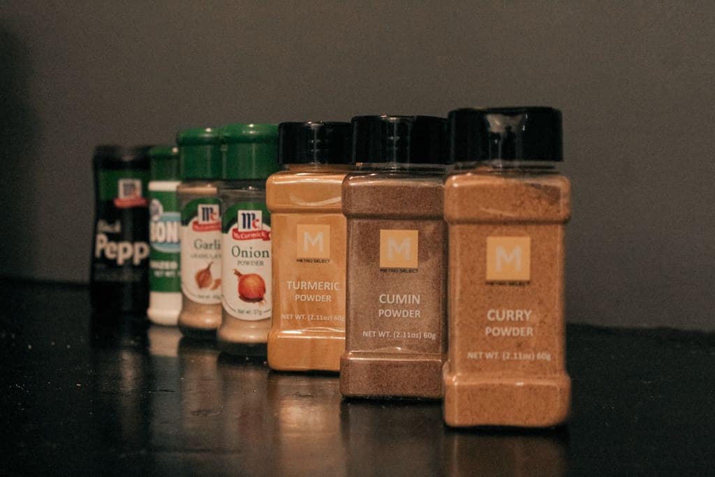 A collection of spice containers is arranged on a dark surface. The spices include black pepper, garlic powder, onion powder, turmeric, cumin, and curry powder, each in clear jars with labeled tops, providing a range of cooking options.