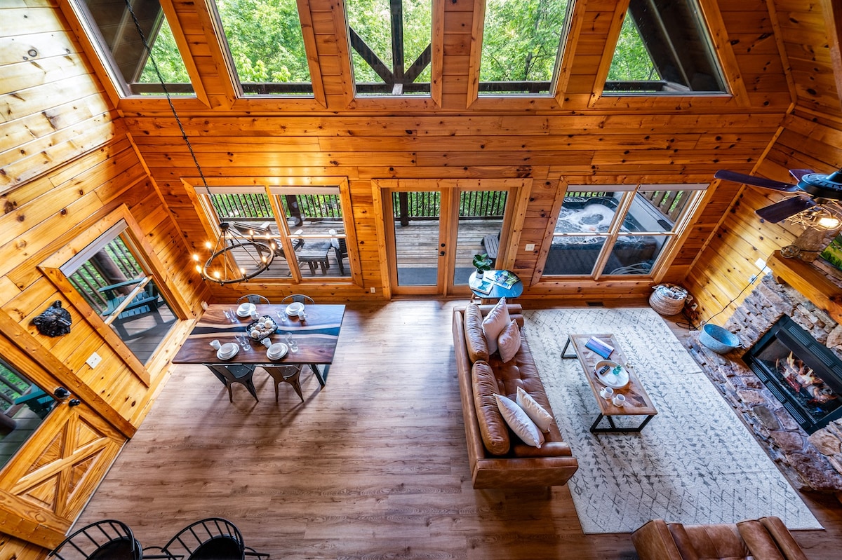 An airy interior is highlighted by wooden walls and a vaulted ceiling with exposed beams. A dining table is set for four adjacent to a comfortable seating area, which features a leather sofa and decorative accents. Large windows provide views of the surrounding treetops.