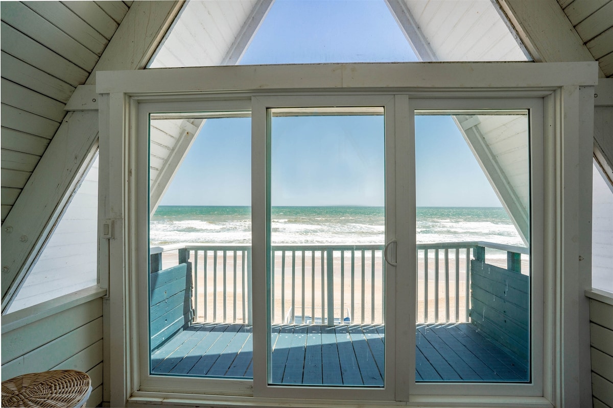 A large sliding glass door is framed by an A-frame structure, showcasing stunning ocean views. Light enters from both sides, illuminating a wooden deck that overlooks the beach and waves. The scene captures the tranquil beauty of the coastal environment.