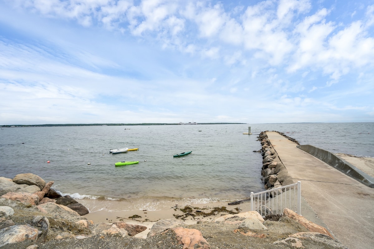 A serene waterfront view features gentle waves lapping against a sandy shore, with a rocky jetty extending into the water. Colorful kayaks are moored nearby, and a cloudy sky adds depth to the tranquil scene.
