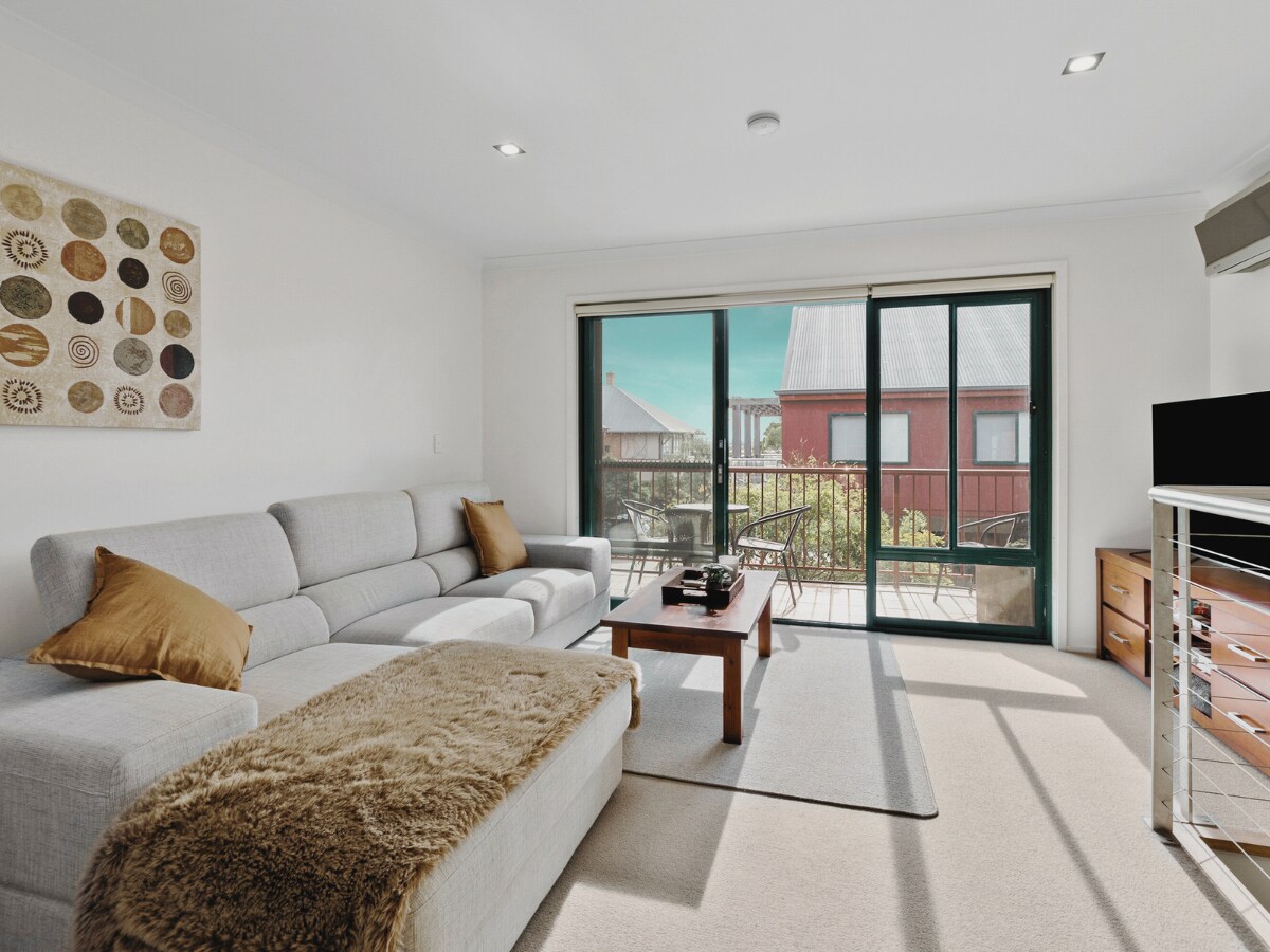 A bright living area features a spacious light grey sectional sofa complemented by decorative cushions. A wooden coffee table sits centrally, and sunlight filters through large sliding glass doors, leading to a balcony. Soft beige carpeting enhances the room's cozy ambience.