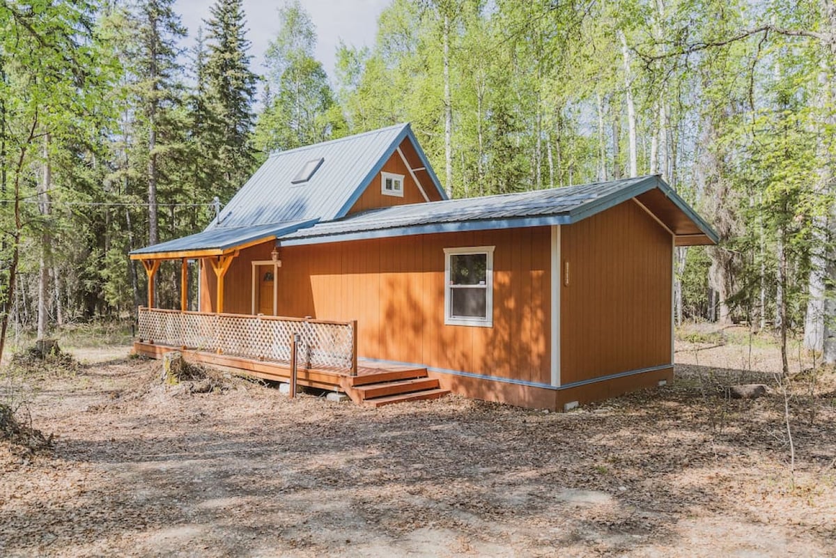 A cabin exterior is shown, featuring warm orange wood siding and a metal roof. A wraparound porch with wooden railings offers accessibility, surrounded by trees and natural landscape, highlighting a serene outdoor setting.