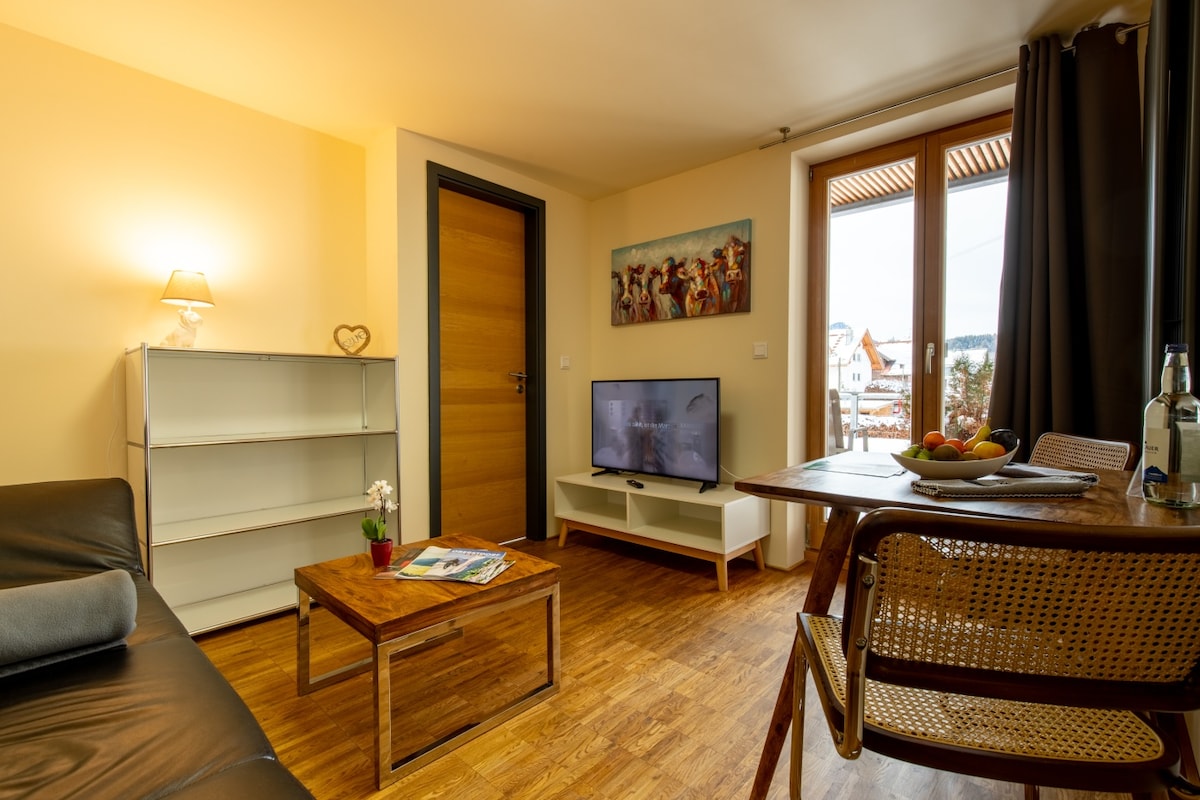 A cozy living area features a black leather sofa and a wooden coffee table, complemented by a dining table with woven chairs. A flat-screen TV is positioned on a white console. Natural light enters through the balcony doors, providing a view of the surroundings.