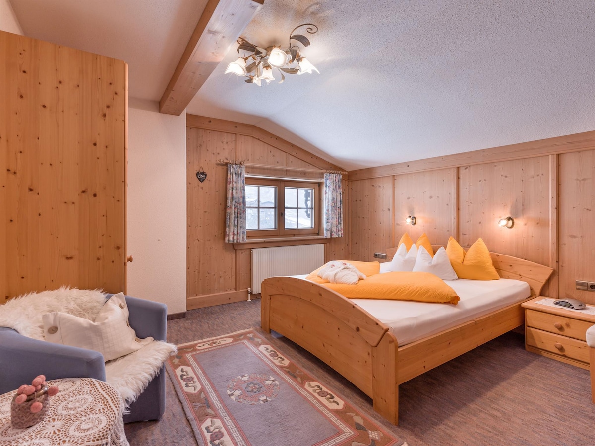 A warm, wooden-paneled bedroom features a spacious bed with orange accent pillows. A cozy armchair is positioned nearby, and decorative curtains frame a window that lets in natural light. A patterned area rug adds texture to the floor.
