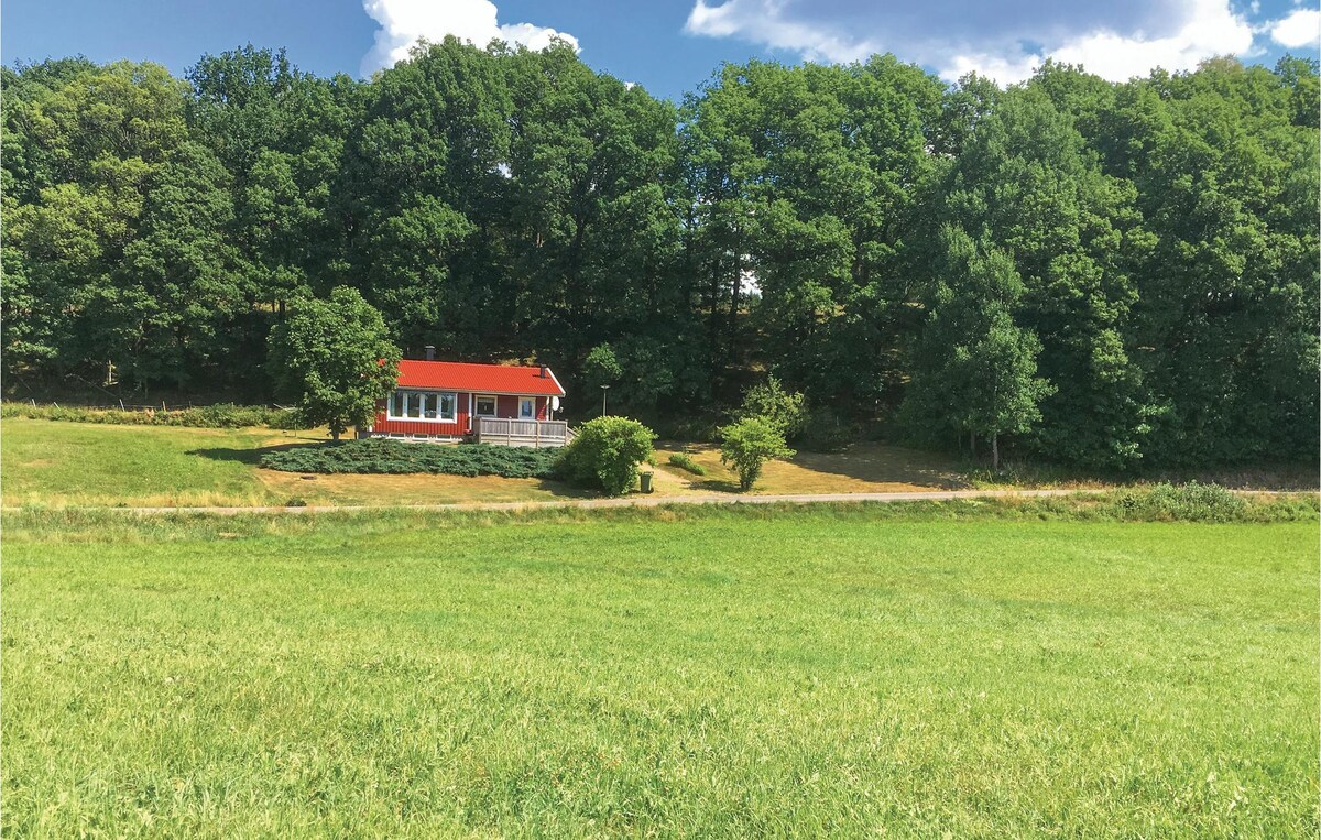 A charming cottage with a red roof is nestled among lush trees, set against a backdrop of expansive green meadows. The structure features a welcoming porch that overlooks the landscape, providing a serene atmosphere in a natural setting.