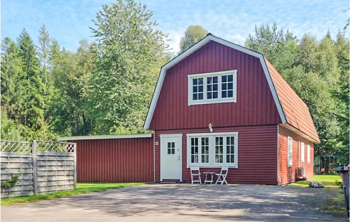 A charming red house with a steeply pitched roof is surrounded by lush greenery. A simple patio area features two white chairs and a small table, enhancing the inviting exterior. The driveway leads to a garage, contributing to the welcoming appearance of the property.