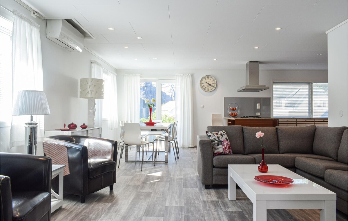 A spacious living area features a comfortable brown sofa, contrasting black armchairs, and a light-colored coffee table. Natural light filters through large windows, illuminating a dining table and a well-equipped kitchen in the background. Simple decor pieces add a touch of color.