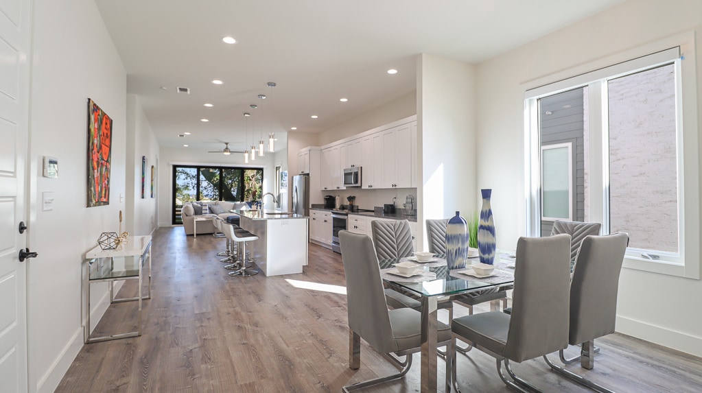 A contemporary open-concept living space features a dining area with a glass table surrounded by six chairs. A modern kitchen with white cabinetry and stainless steel appliances is visible, alongside large windows that bring in natural light and offer views of the outdoors.