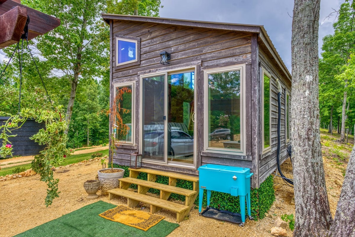 A tiny home exterior is showcased amidst lush greenery, featuring large glass windows that invite natural light. Steps lead to a welcoming entrance, adorned with a small mat. A bright blue cooler sits to the right, enhancing the charming outdoor atmosphere.